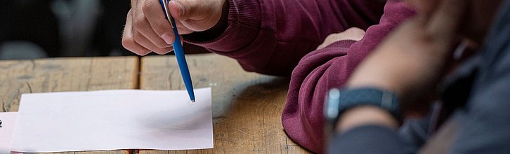 Hand hält Stift, der auf Papier zeigt. Eine andere Person schaut zu.